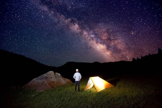 A woman, endurance athlete Meghan Hicks, looks up at the Milky Way and night sky next to her tent while camping in Big Meadow on her fastpack of the Tahoe Rim Trail, CA.